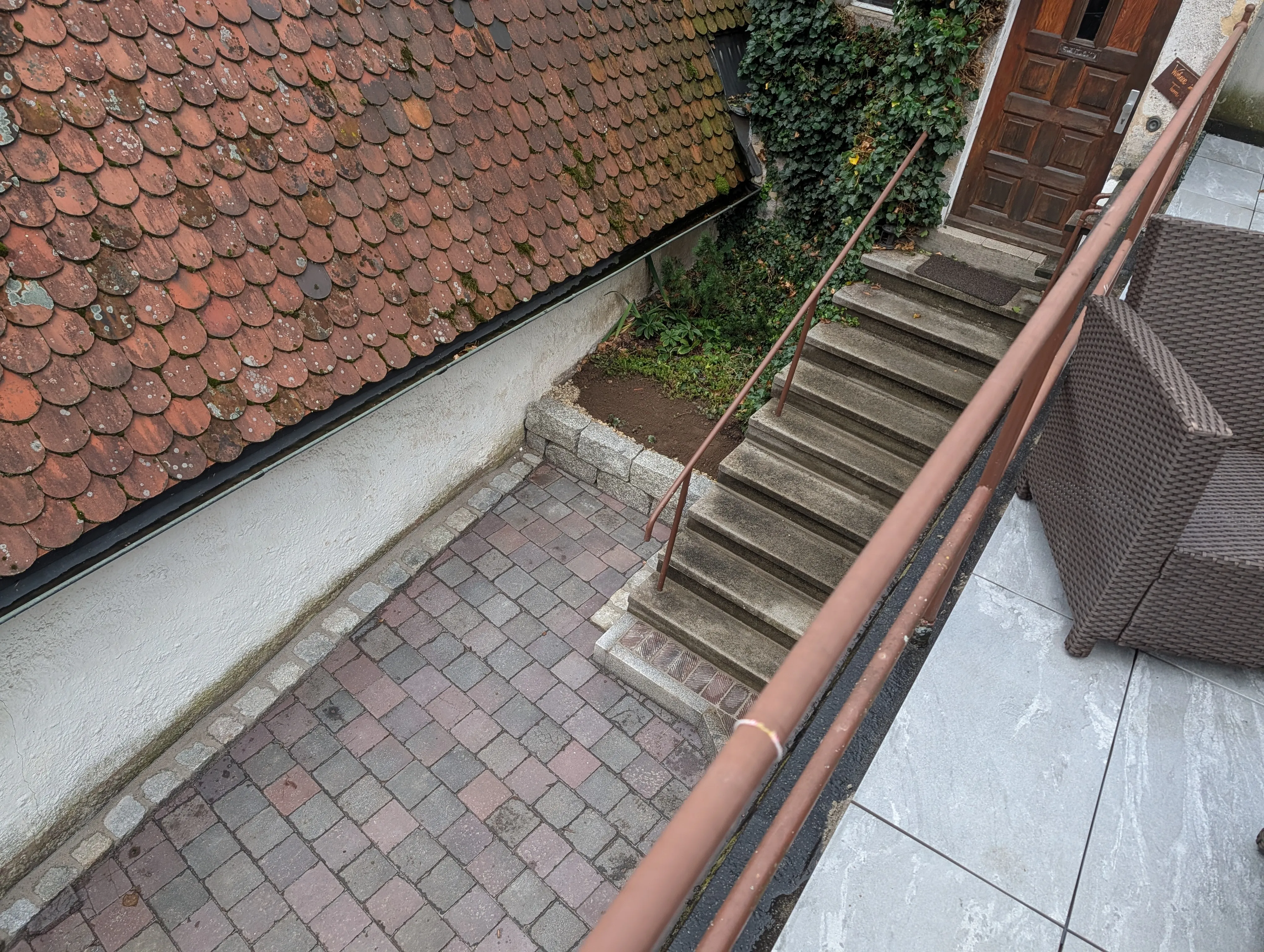 Von der Terrasse hinunter auf den gepflasterten Hof, die Treppe zur Haustür und das Biberschwanzdach nebenan.