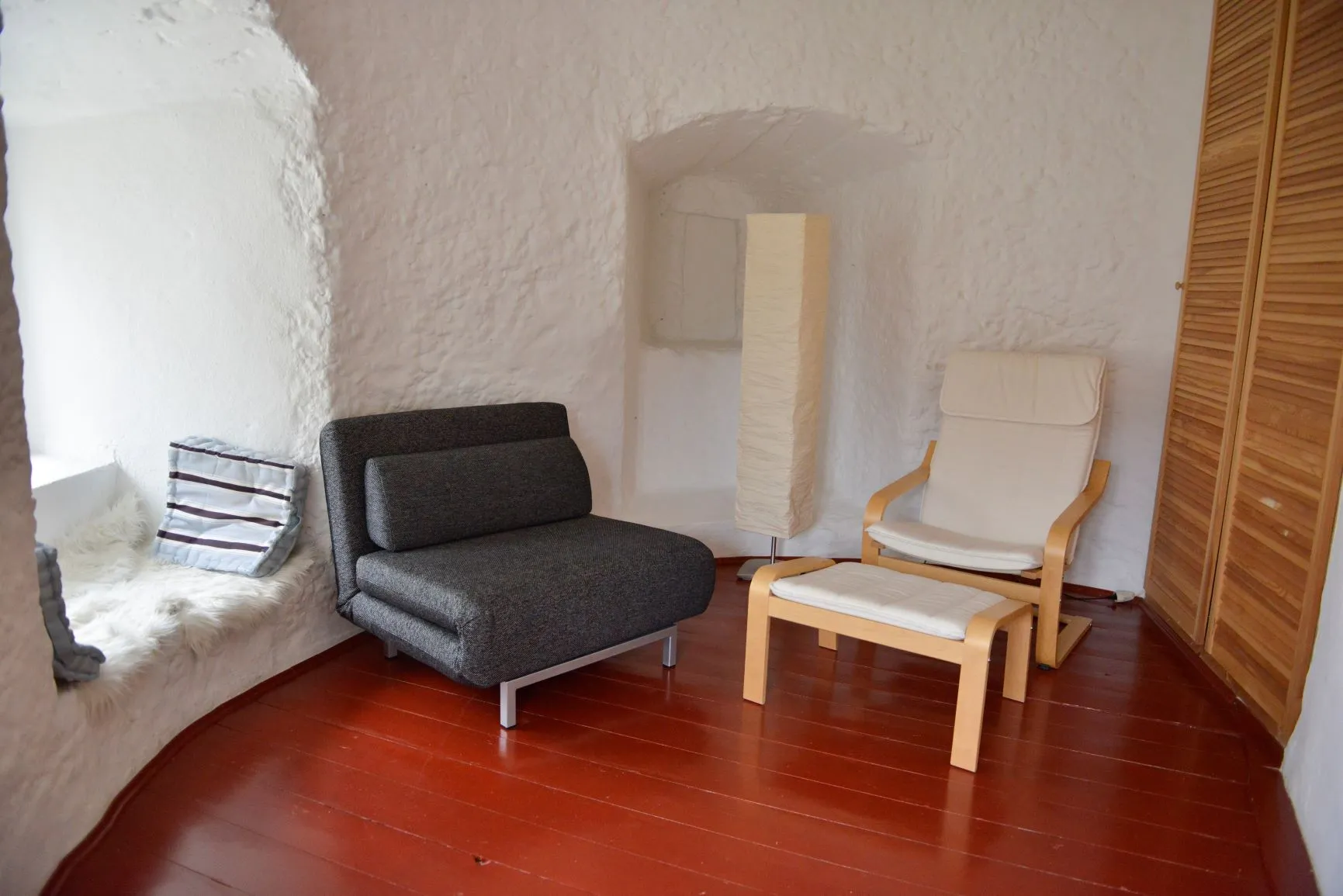Die Lounge im Mittelgeschoss: Fenstersitz mit Schaffell und Kissen, grauer Schlafsessel, heller Sessel mit Hocker, eine Stehlampe in der Mauernische und ein Schrank mit Lamellentüren, auf rotem Dielenboden.