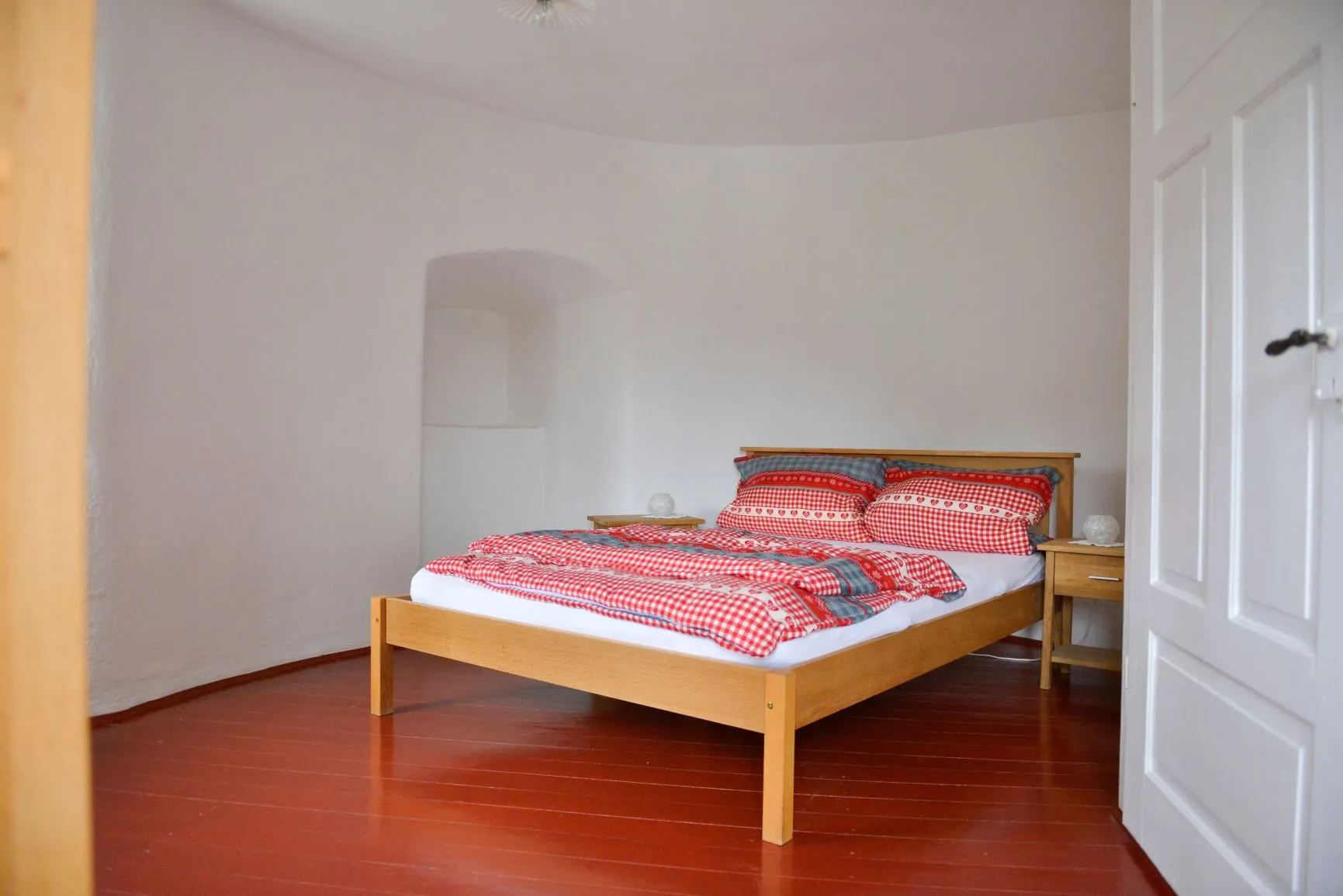 Das Schlafzimmer im Obergeschoss: Doppelbett mit rot-weiß karierter Bettwäsche, zwei Nachttische mit Lampen, eine Nische in der gerundeten Wand, roter Dielenboden.